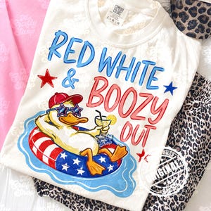 Pode incluir: Camiseta creme com a frase "RED WHITE & BOOZY OUT" em azul e vermelho. Um pato de desenho animado usando óculos de sol e chapéu flutua em uma boia vermelha, branca e azul, segurando um coquetel.