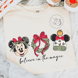 May include: Cream-colored sweatshirt featuring three Christmas-themed designs: Minnie Mouse with a bow, a wreath with a red bow, and a Mickey Mouse hat. The phrase "believe in the magic" is written in cursive below the designs.