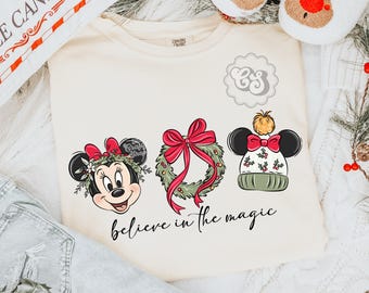 May include: Cream-colored sweatshirt featuring three Christmas-themed designs: Minnie Mouse with a bow, a wreath with a red bow, and a Mickey Mouse hat. The phrase "believe in the magic" is written in cursive below the designs.