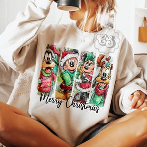 Puede incluir: Sudadera color crema con un dise&ntilde;o festivo con personajes de dibujos animados. El dise&ntilde;o incluye a Goofy, Mickey Mouse, Minnie Mouse y Pluto, cada uno con atuendos navide&ntilde;os. Las palabras "Merry Christmas" est&aacute;n escritas en una fuente cursiva.
