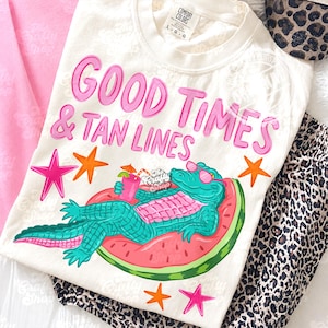 Może przedstawiać: Kremowa koszulka z różowym napisem "GOOD TIMES & TAN LINES". Koszulka przedstawia turkusowego aligatora w okularach przeciwsłonecznych, relaksującego się na pływaku w kształcie arbuza, trzymającego drinka. Różowe i pomarańczowe gwiazdki dopełniają projekt.