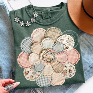 May include: A sage green sweatshirt with a large, appliqued flower design. The flower is made of various fabric patches in pastel colors and floral patterns. A silver necklace with flower pendants and a brown hat are also visible.