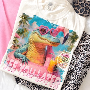 May include: Cream-colored t-shirt featuring a colorful graphic of an alligator wearing heart-shaped sunglasses and floating on a flamingo floatie. The text "See Ya Later" is below the alligator. The shirt has a Comfort Colors tag.