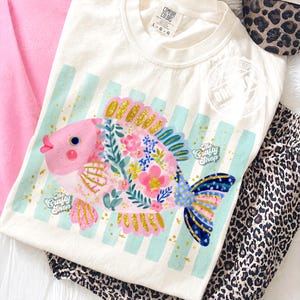May include: Cream-colored t-shirt featuring a colorful fish design. The fish is pink, blue, and gold, with floral details and the text "The Crafty Shop". The shirt has a crew neck and short sleeves. The shirt is laying on a pink surface.