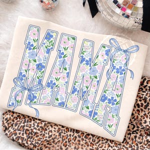 May include: Cream-colored sweatshirt with the word "MAMA" in large, block letters. The letters are filled with a floral pattern in shades of blue, pink, and green. Light blue bows are on the letters. The sweatshirt is folded on a leopard print fabric.