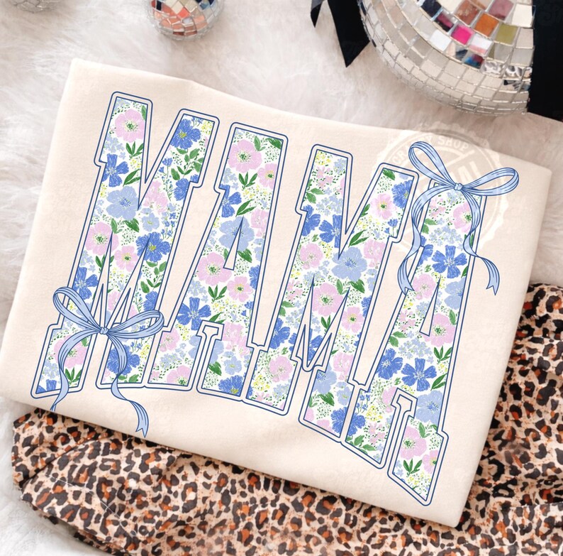 May include: Cream-colored sweatshirt with the word "MAMA" in large, block letters. The letters are filled with a floral pattern in shades of blue, pink, and green. Light blue bows are on the letters. The sweatshirt is folded on a leopard print fabric.
