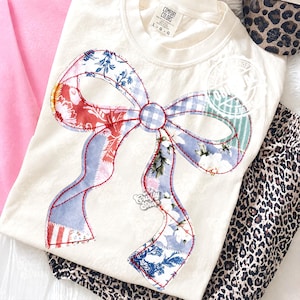 Pode incluir: Camiseta creme com um grande design de laço de patchwork. O laço apresenta vários tecidos florais e estampados em vermelho, azul e branco, delineados com costuras vermelhas. A camiseta está dobrada com outras roupas.
