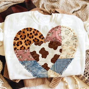 May include: White sweatshirt with a patchwork heart design. The heart is made from different fabrics, including leopard print, floral patterns, denim, and cowhide. The design is stitched onto the front of the garment.