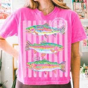 May include: A vibrant pink t-shirt with three rainbow trout designs. The fish are depicted with colourful scales, set against pink and white striped background. The shirt includes the text "The Crafty Shop Original".