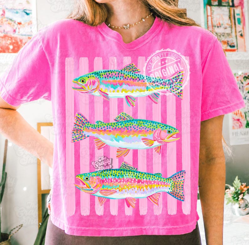 May include: A vibrant pink t-shirt with three rainbow trout designs. The fish are depicted with colourful scales, set against pink and white striped background. The shirt includes the text "The Crafty Shop Original".