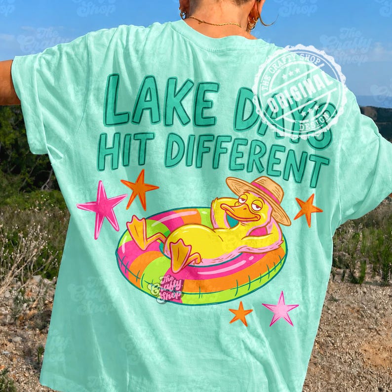 May include: A light teal t-shirt with the text "LAKE DAYS HIT DIFFERENT" in teal. The shirt features a cartoon duck wearing a hat and floating on a colourful inflatable ring. Pink and orange starfish are also visible.