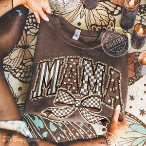 May include: A brown sweatshirt with the word "MAMA" in a decorative font, featuring a bow graphic. The letters have a mix of patterns, including stripes, polka dots, and checkered designs. The sweatshirt is held by hands.