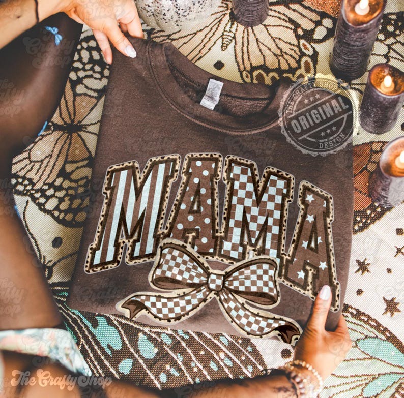 May include: A brown sweatshirt with the word "MAMA" in a decorative font, featuring a bow graphic. The letters have a mix of patterns, including stripes, polka dots, and checkered designs. The sweatshirt is held by hands.