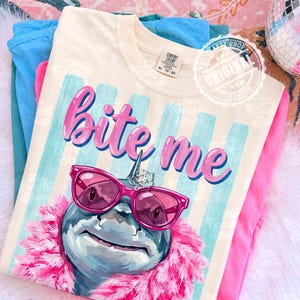 K&ouml;nnte beinhalten: Cremefarbenes T-Shirt mit dem Schriftzug "Bite Me" in rosa Schrift. Das Shirt zeigt einen Hai mit rosa Sonnenbrille und rosa Boa. Der Hintergrund hat blau-wei&szlig;e Streifen.