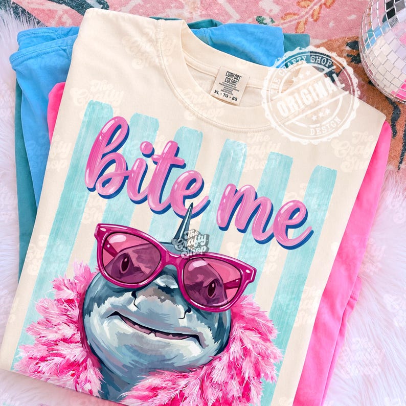 K&ouml;nnte beinhalten: Cremefarbenes T-Shirt mit dem Schriftzug "Bite Me" in rosa Schrift. Das Shirt zeigt einen Hai mit rosa Sonnenbrille und rosa Boa. Der Hintergrund hat blau-wei&szlig;e Streifen.