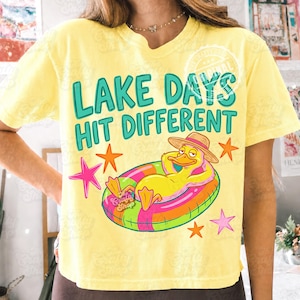 May include: Yellow t-shirt with the text "LAKE DAYS HIT DIFFERENT" in teal. A cartoon duck wearing a straw hat and floating on a colourful inflatable ring is featured. Pink and orange stars surround the duck.
