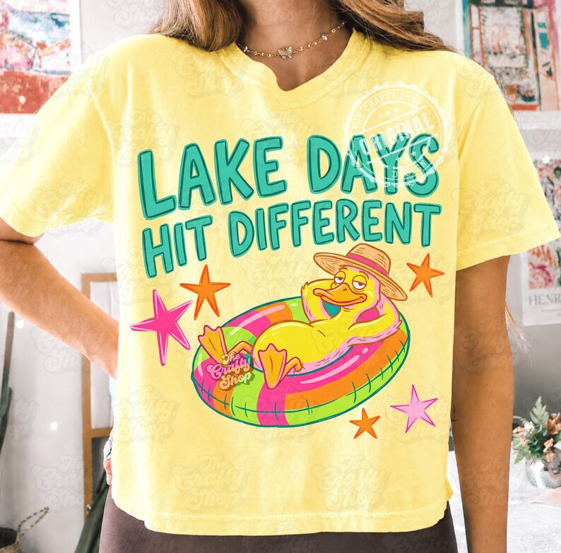 May include: Yellow t-shirt with the text "LAKE DAYS HIT DIFFERENT" in teal. A cartoon duck wearing a straw hat and floating on a colourful inflatable ring is featured. Pink and orange stars surround the duck.