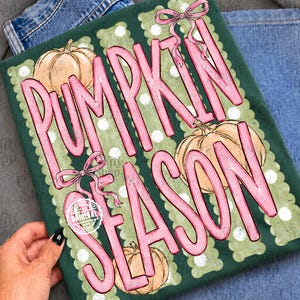 May include: A dark green t-shirt featuring a graphic print with the text PUMPKIN SEASON in large pink letters. The design includes three light orange pumpkins, pink bows, and a background pattern of vertical scalloped stripes with white dots.
