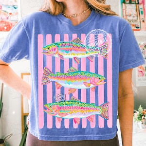 May include: Blue t-shirt with a graphic of three rainbow trout in vibrant colours. The fish are set against pink vertical stripes. The shirt has the text "The Crafty Shop Original".
