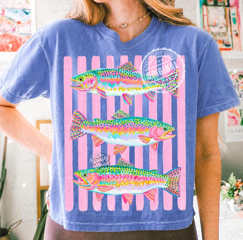 May include: Blue t-shirt with a graphic of three rainbow trout in vibrant colours. The fish are set against pink vertical stripes. The shirt has the text "The Crafty Shop Original".