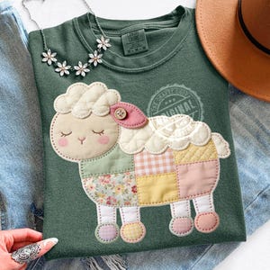 Pode incluir: Camisola verde Comfort Colors com um design de ovelha em patchwork. A ovelha tem uma cabeça creme, bochechas rosa e um detalhe de botão. O corpo em patchwork inclui blocos florais, xadrez e de cor lisa. Um colar floral e um chapéu castanho também são visíveis.