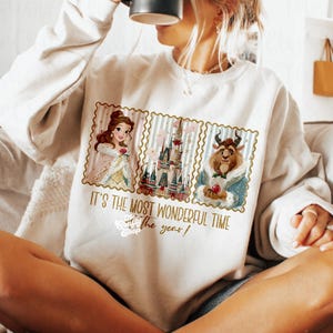 Puede incluir: Sudadera color crema con un gr&aacute;fico de Bella, la Bestia y un castillo. El dise&ntilde;o est&aacute; enmarcado con un borde de sello postal e incluye el texto "IT'S THE MOST WONDERFUL TIME of the year!"