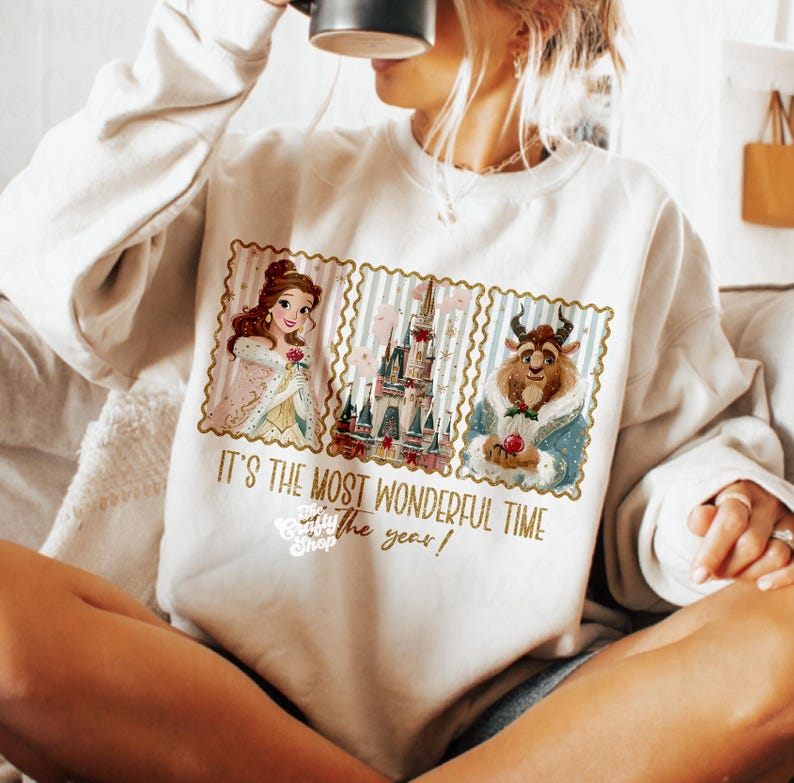 Puede incluir: Sudadera color crema con un gr&aacute;fico de Bella, la Bestia y un castillo. El dise&ntilde;o est&aacute; enmarcado con un borde de sello postal e incluye el texto "IT'S THE MOST WONDERFUL TIME of the year!"