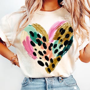 Pode incluir: Camiseta creme com um design de coração colorido. O coração é composto por pinceladas em rosa, dourado, verde-azulado e manchas pretas. A camiseta tem mangas curtas e é combinada com jeans azuis.
