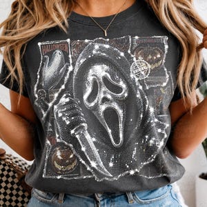 May include: A charcoal gray t-shirt featuring a graphic print of a masked figure holding a knife, surrounded by smaller images of pumpkins and corn. The design has a shimmering, mosaic-like texture and includes the text "HALLOWEEN" and "ORIGINAL".