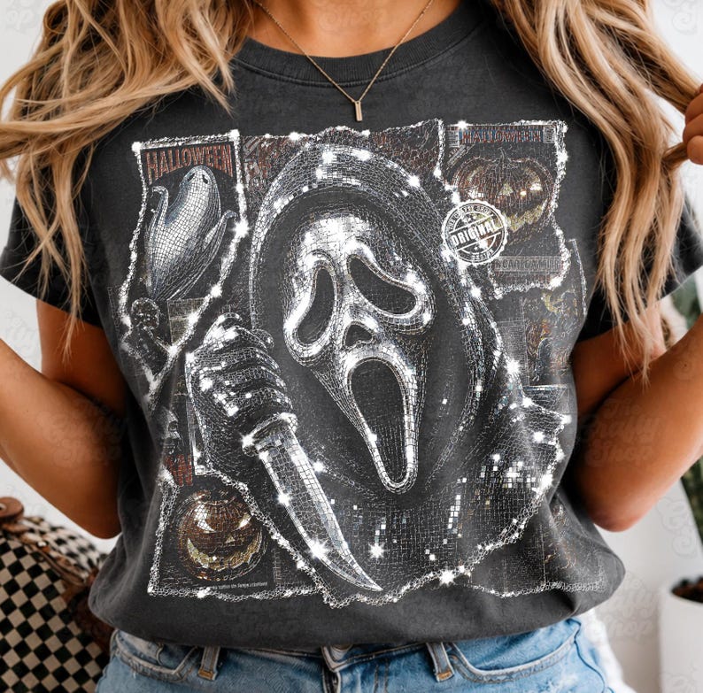 May include: A charcoal gray t-shirt featuring a graphic print of a masked figure holding a knife, surrounded by smaller images of pumpkins and corn. The design has a shimmering, mosaic-like texture and includes the text "HALLOWEEN" and "ORIGINAL".