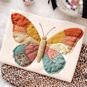 Pode incluir: Uma almofada creme com um grande design de borboleta em patchwork. A borboleta apresenta vários retalhos de tecido em tons de laranja, amarelo, verde-azulado e rosa. O centro da borboleta tem uma textura semelhante à juta, e as asas são costuradas.