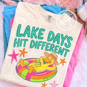 May include: A cream-coloured t-shirt with the text "LAKE DAYS HIT DIFFERENT" in teal. The shirt features a cartoon duck wearing a hat and floating in a colourful inflatable ring. Pink, orange, and teal stars are also on the shirt.
