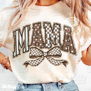 May include: A cream-coloured t-shirt featuring the word "MAMA" in a brown, blue, and white patterned design. The letters have stripes, polka dots, and stars. A checkered bow is below the word. This is a casual wear item.