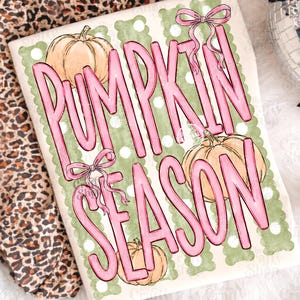 May include: A white t-shirt featuring a graphic print with the text PUMPKIN SEASON in large pink letters. The design includes three light orange pumpkins, two pink bows, and a background of vertical sage green stripes with white polka dots.