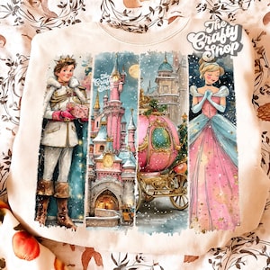 Pode incluir: Camisola creme com um gráfico colorido de um príncipe, uma princesa e um castelo. O gráfico inclui uma carruagem rosa, um castelo e o texto "The Crafty Shop". O design tem um tema de inverno.