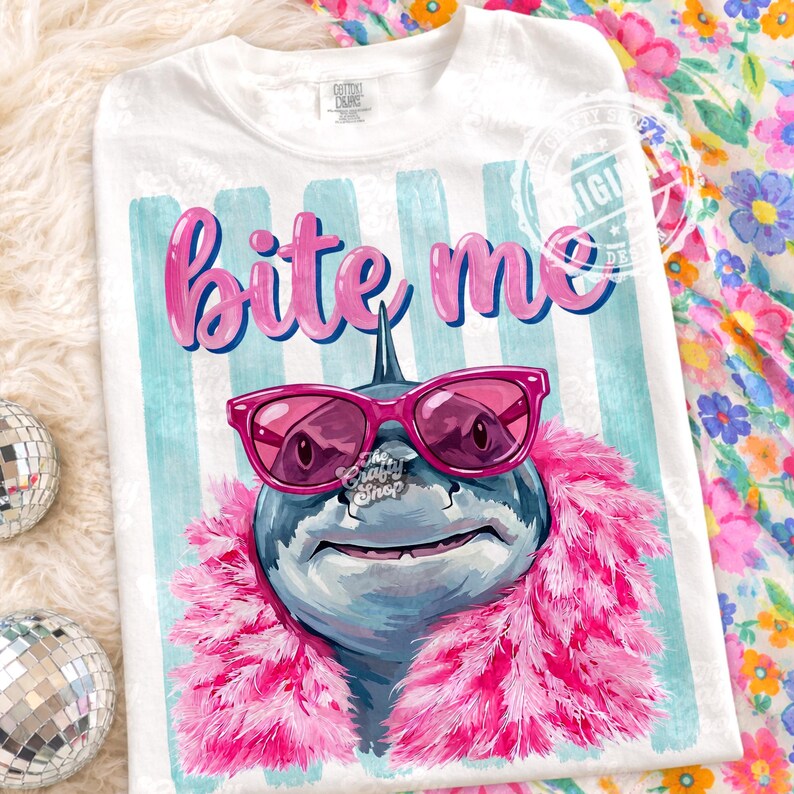 K&ouml;nnte beinhalten: Wei&szlig;es T-Shirt mit einer Grafik eines Hais mit rosa Sonnenbrille und einer rosa Boa. Der Text "bite me" steht in rosa Buchstaben &uuml;ber dem Hai. Der Hintergrund hat blau-wei&szlig;e Streifen.