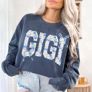 May include: A navy blue sweatshirt with the word "GIGI" in large, floral-patterned letters. The letters are filled with blue, pink, and white flowers. Light blue bows are on the sides of the letters. The sweatshirt has long sleeves.