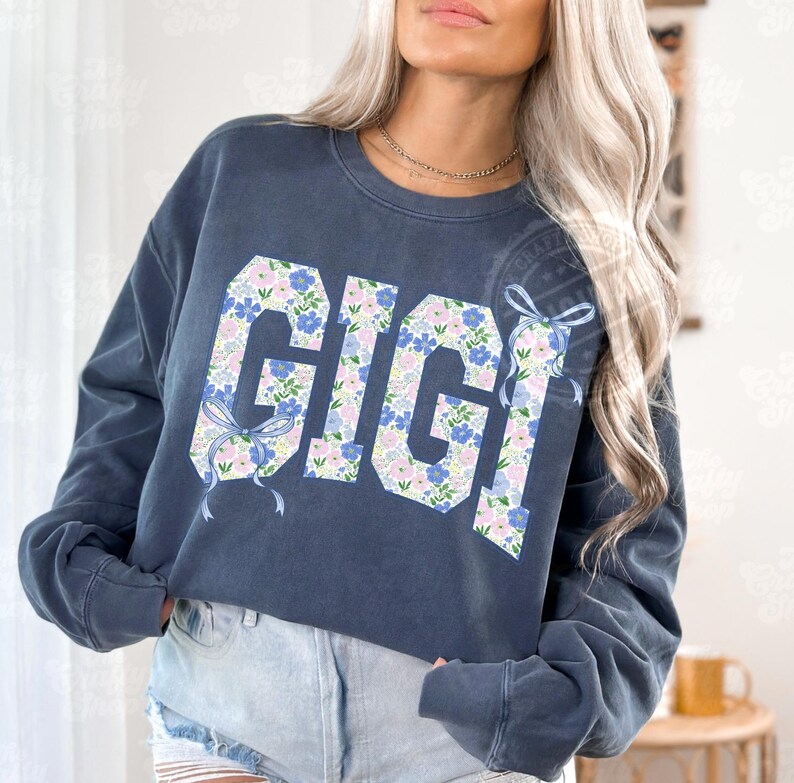 May include: A navy blue sweatshirt with the word "GIGI" in large, floral-patterned letters. The letters are filled with blue, pink, and white flowers. Light blue bows are on the sides of the letters. The sweatshirt has long sleeves.