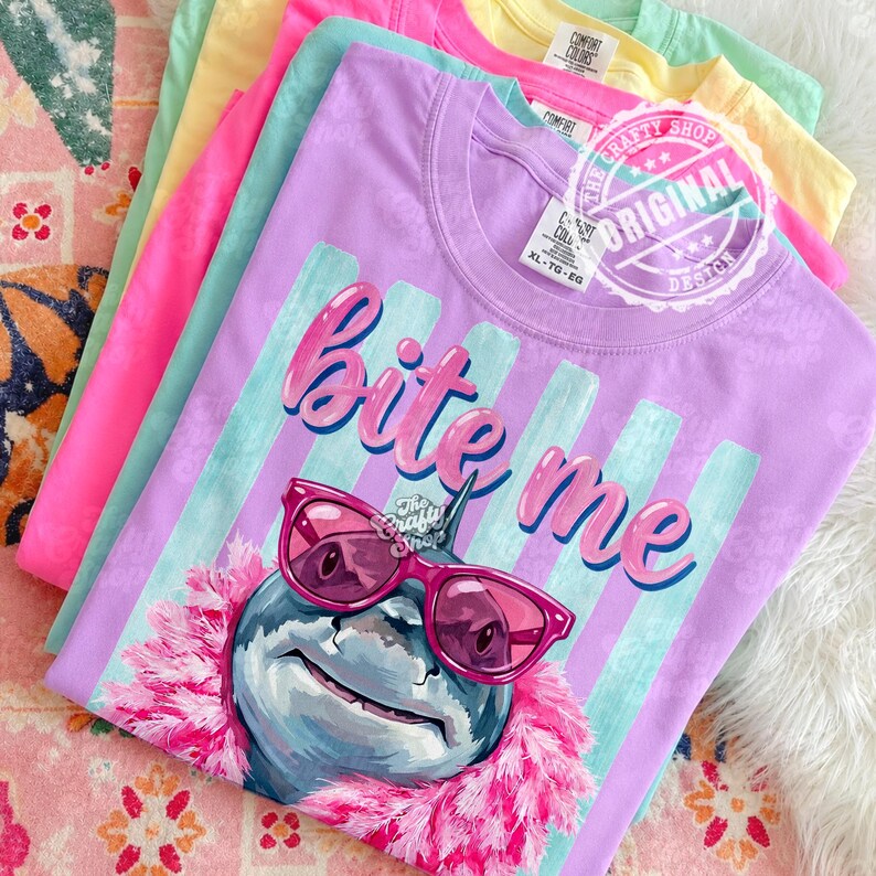 K&ouml;nnte beinhalten: Ein Stapel farbenfroher T-Shirts in Rosa, Lila, Gelb und Gr&uuml;n. Das vordere Shirt ist lila mit einer Hai-Grafik mit rosa Sonnenbrille und den Worten "Bite Me". Die Shirts haben das Etikett "The Crafty Shop".