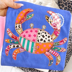 Pode incluir: Uma camiseta azul com um design de caranguejo colorido. O caranguejo é feito de vários tecidos estampados, incluindo estrelas rosa, bolinhas laranja e estampa de leopardo. As garras e pernas do caranguejo são adornadas com lantejoulas. A camiseta tem um logotipo "The Crafty Shop Original".