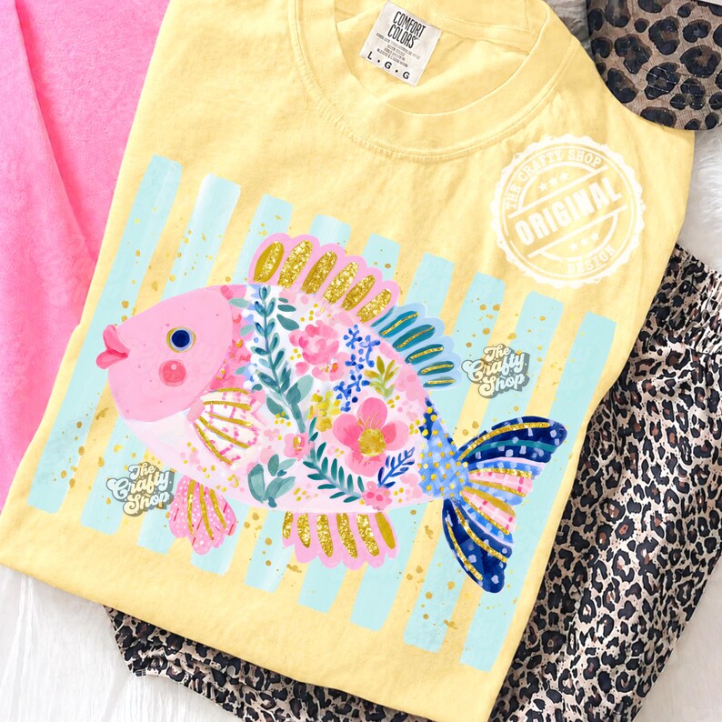 May include: Yellow t-shirt with a colorful fish graphic. The fish features pink, gold, and blue details, with floral accents. The shirt has light blue vertical stripes and the text "The Crafty Shop".