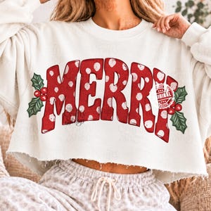 Pode incluir: Uma camisola curta de cor creme com o texto vermelho MERRY com bolinhas brancas. O design é acentuado por folhas de azevinho verdes e bagas vermelhas em ambos os lados. A camisola tem uma bainha crua e inacabada.