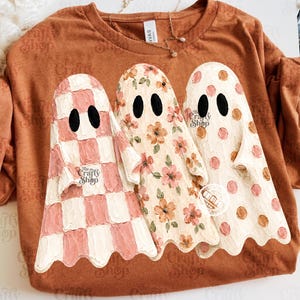 Pode incluir: Uma t-shirt cor de ferrugem com um gráfico de três fantasmas com olhos ovais pretos. O primeiro fantasma tem um padrão xadrez rosa e branco, o segundo uma estampa floral e o terceiro bolinhas rosas e marrons. O logótipo The Crafty Shop é visível.