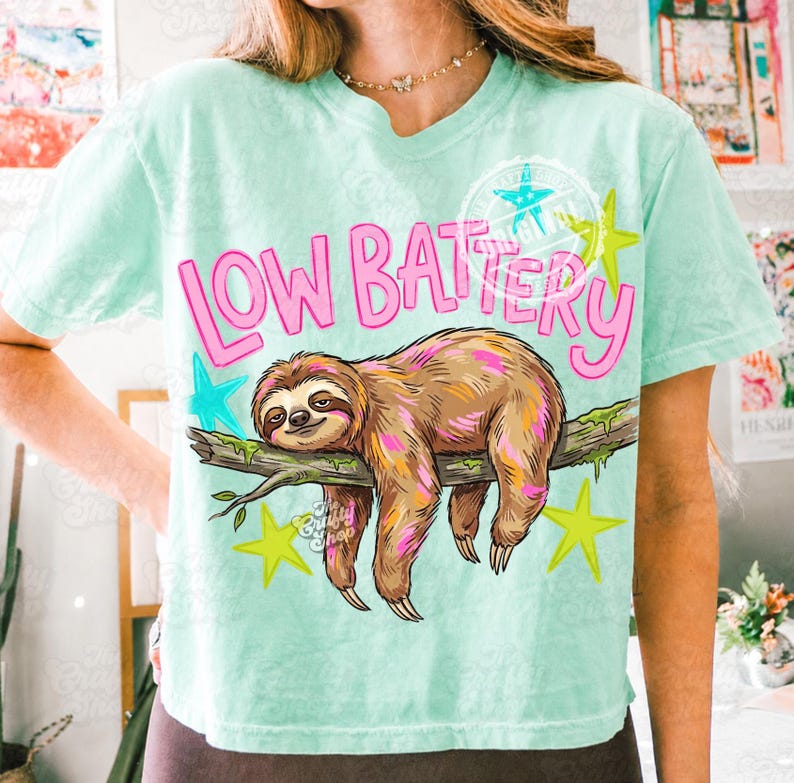 Puede incluir: Camiseta verde claro con las palabras "LOW BATTERY" en rosa, con un perezoso de dibujos animados descansando en una rama. La camiseta tiene un corte holgado y est&aacute; decorada con estrellas.