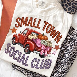 Pode incluir: Uma t-shirt cor de creme com um gráfico de uma carrinha vermelha a transportar abóboras, um chapéu de cowboy cor-de-rosa e um laço cor-de-rosa. O texto diz SMALL TOWN SOCIAL CLUB em letras castanhas e roxas, com duas estrelas cor de laranja.