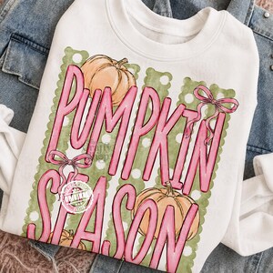 May include: A white crewneck sweatshirt featuring a graphic print with the text PUMPKIN SEASON in large pink letters. The design includes two orange pumpkins, a green scalloped background with white polka dots, and two pink bows.