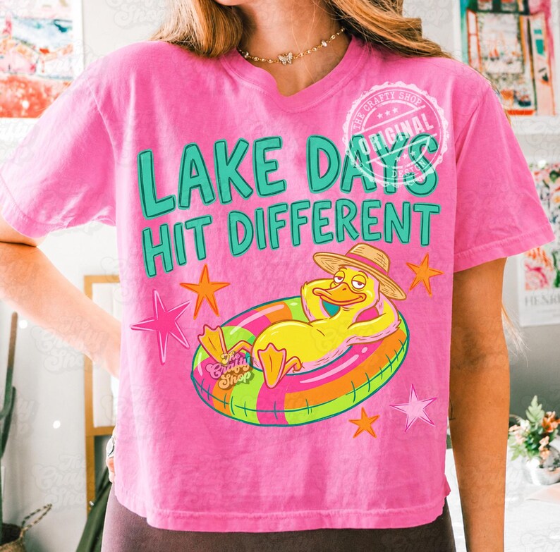 May include: A pink t-shirt with the slogan "LAKE DAYS HIT DIFFERENT" in green. The design includes a cartoon duck in a straw hat on a colourful inflatable ring, with orange and pink star graphics. The shirt is a casual summer style.