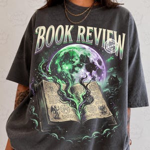 Pode incluir: Uma t-shirt cinzenta-carvão de corte oversize com o texto BOOK REVIEW em letra negrito. O gráfico mostra um livro de feitiços aberto com fumo mágico verde a subir para uma grande lua roxa e verde, junto à silhueta de uma casa.