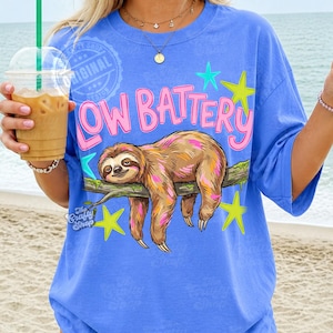 Puede incluir: Camiseta azul con el texto "LOW BATTERY" en rosa, con un perezoso de dibujos animados en una rama. El perezoso tiene pelaje marr&oacute;n con detalles rosas y amarillos, rodeado de estrellas verdes y amarillas.