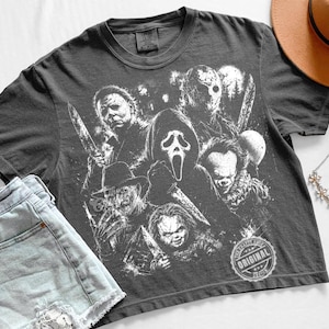 May include: A charcoal gray short-sleeve t-shirt featuring a white graphic print of five horror movie characters, including a masked figure with a knife, a hockey-masked killer, a clown, a fedora-wearing villain, and a small doll-like figure.